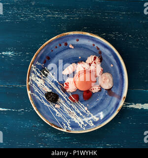 Tasty sweet dessert consisted of pink macaron, meringues, raspberries, blackberries and whipped cream lying on flat plate standing on blue old wooden  Stock Photo