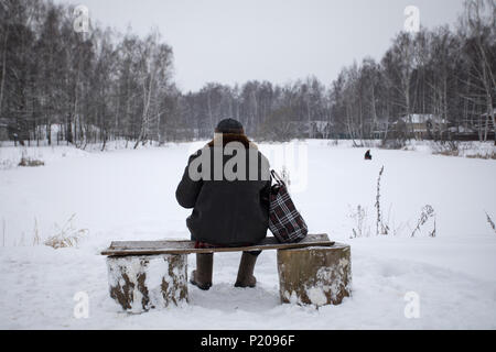 Old man sits on a bench in the winter in front of a frozen pond with fishermen, the view from the back. Stock Photo