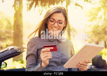 Smiling woman standing by her new car making online payment on her tablet computer outside on a sunny summer day Stock Photo