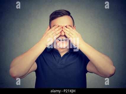 Young cheerful man covering eyes with hands anticipating surprise while posing on gray backdrop. Stock Photo