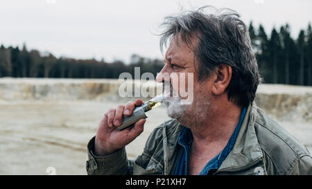 Old man with casual outfit is smoking e-cigarette in forest area Stock Photo