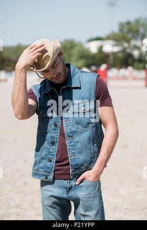 young handsome man wearing cowboy hat and denim vest standing on ranch Stock Photo