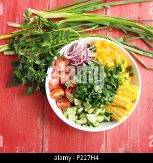White plate with cut vegetables for a vegan salad on the red wooden table. Colorful photo. Corn, cherry tomatoes, bell pepper, onion, cucumber and gar Stock Photo