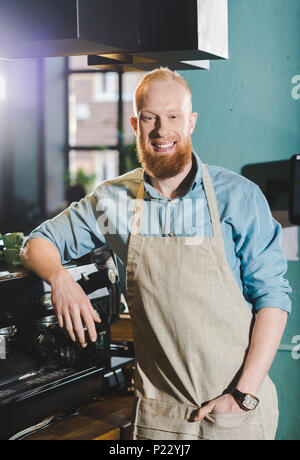 young bearded smiling male barista in apron standing near coffee machine Stock Photo
