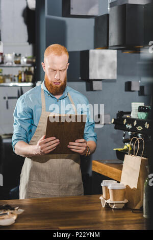 serious bearded barista in apron holding clipboard while working in coffee shop Stock Photo