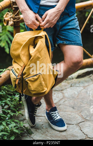 cropped shot of stylish man with yellow backpack leaning on fence in park Stock Photo
