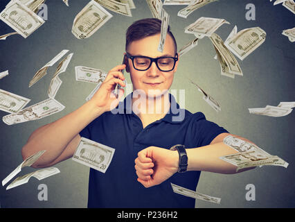 Busy businessman looking at wrist watch, talking on mobile phone under cash rain. Time is money concept Stock Photo