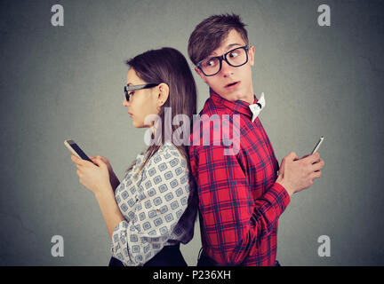 Young couple standing back to back, holding smartphones. Jealous man looking over his shoulder at his girlfriend phone trying to see what she is texti Stock Photo