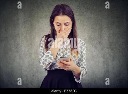 Young woman covering mouth in astonishment while reading message on smartphone looking stunned. Stock Photo