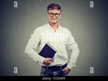 Young cheerful man in glasses holding book and looking happily at camera excited with studies. Stock Photo