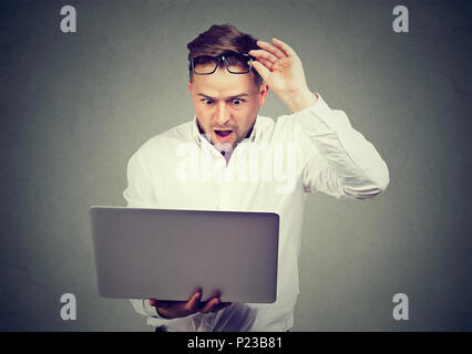 Surprised young man lifting up eyeglasses while watching laptop in amazement. Stock Photo