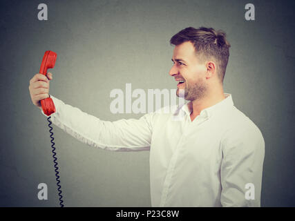 Laughing casual man in white shirt holding red receiver of phone in outstretched hand on gray background. Stock Photo