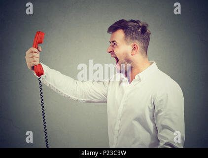 Young man in white shirt holding red phone handset and screaming in anxiety on gray background. Stock Photo