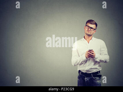 Handsome man in glasses holding phone in hands and looking up in contemplation on gray backdrop. Stock Photo