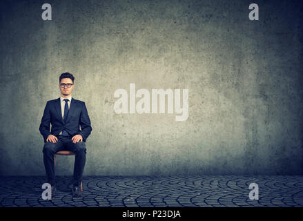 Confident man in suit and glasses sitting on chair waiting for job interview on gray background. Stock Photo
