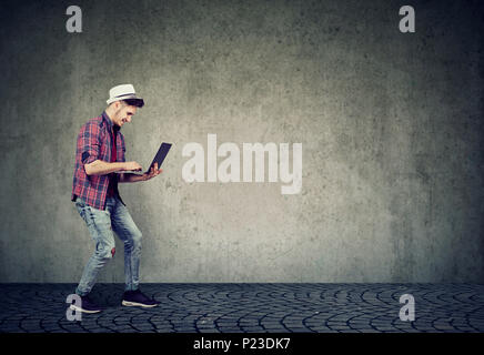 Side view of stylish hipster man in hat holding and surfing laptop standing on gray background. Stock Photo