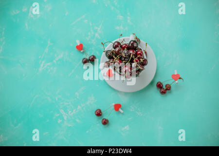Red ripe cherries in a white cup isolated on pastel green background. Red heart and sweet berries. Summer composition, copy space.Delicious food for health vegan life with fresh berries Stock Photo