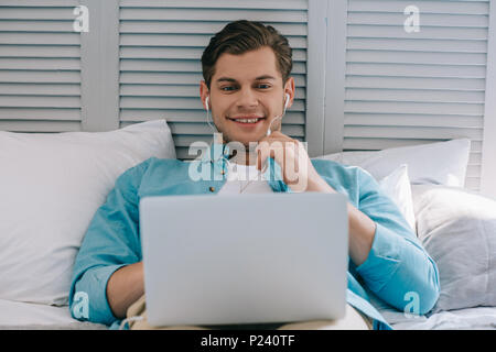 Young man in earbuds using laptop while lying on bed Stock Photo