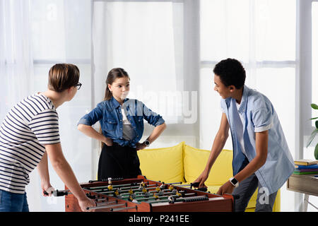 multicultural teen boys playing table football and female friend standing with hands akimbo at home Stock Photo