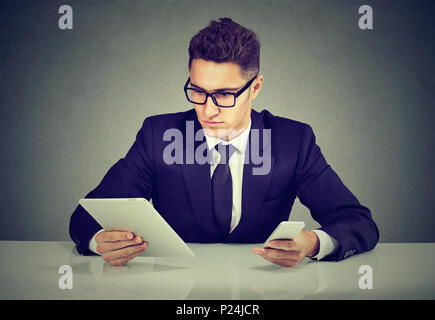Young man in trendy suit posing at desk with smartphone and tablet busy with work. Stock Photo