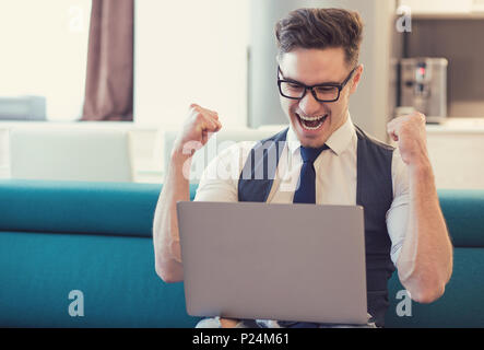 Young man using laptop and celebrating good news looking happy while sitting on couch. Stock Photo