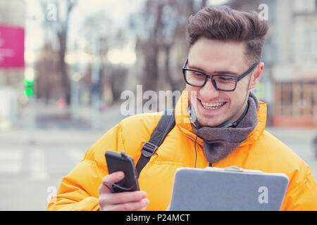 Content happy male student in eyeglasses and headphones messaging via smartphone standing on street. Stock Photo