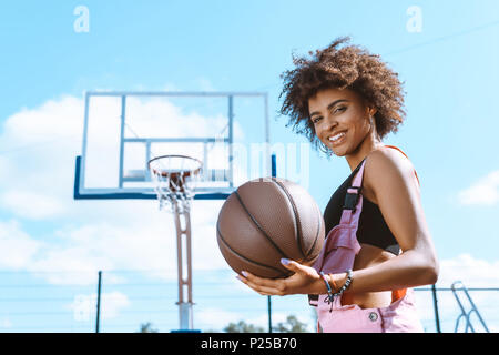african-american woman in sports bra and pink overalls holding a basketball ball at sports court Stock Photo