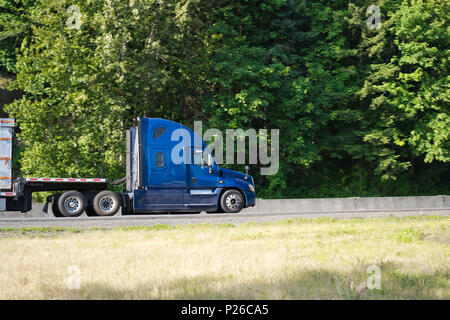Commercial powerful big rig blue long haul semi truck transporting step down semi trailer on the green winding highway with trees on background for de Stock Photo