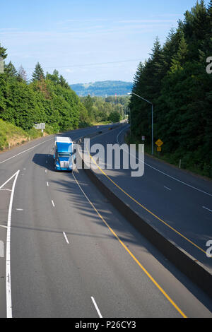 Commercial high profile cab big rig blue long haul semi truck transporting dry van semi trailer on the green winding highway with trees on shoulder si Stock Photo