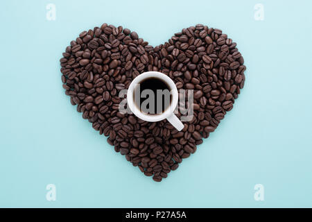 Heart shape made of perfect coffee beans & a cup of black coffee on blue background. Flat lay, minimal. Love coffee espresso concept. Stock Photo