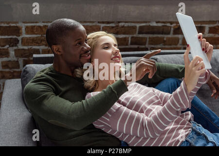 portrait of smiling multicultural couple using tablet together while resting on sofa at home Stock Photo