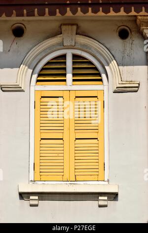 Vertical view of traditional vintage Singapore shop house with antique yellow shutters and arched window on a white wall in historic Little India Stock Photo