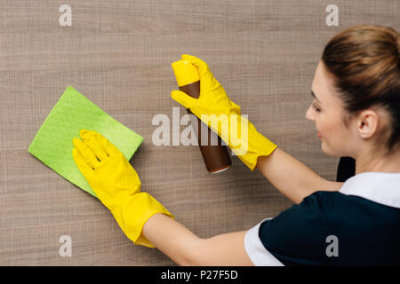 close-up shot of young maid in uniform wiping wood wall with rag and aerosol furniture cleaner Stock Photo