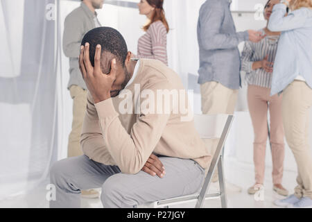 upset african american man sitting on chair while people standing behind during group therapy Stock Photo