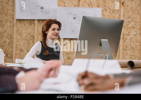 young female architect working with computer at office while colleagues drawong plans on foreground Stock Photo