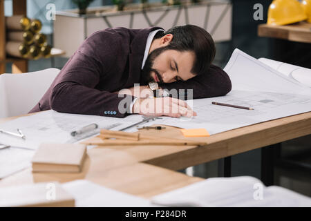 overworked young architect sleeping on building plans at workplace Stock Photo