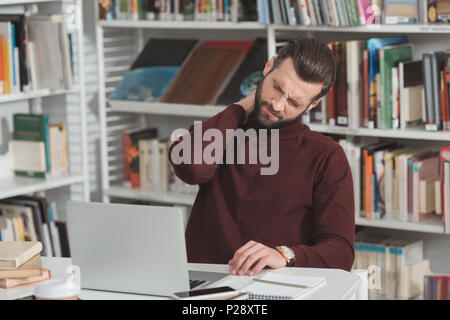 tired student touching neck while working with laptop in library Stock Photo