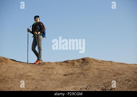 Image of tourist woman in jacket with backpack and walking sticks in background of hill in afternoon Stock Photo