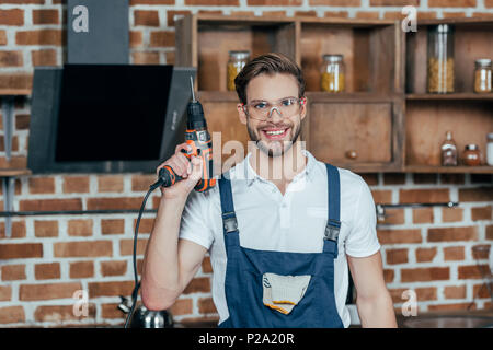 young repairman in protective workwear holding electric drill and smiling at camera Stock Photo