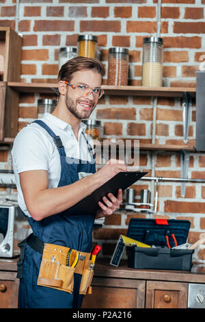handsome young foreman in protective workwear writing on clipboard and smiling at camera Stock Photo