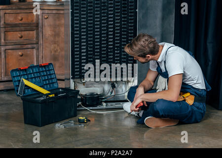 young handyman with toolbox sitting on floor and looking at broken refrigerator Stock Photo