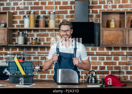 young repairman with wrench and screwdriver smiling at camera while repairing toaster Stock Photo