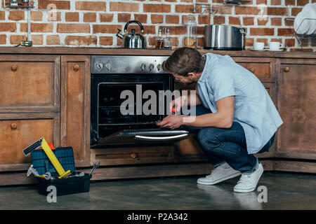 casual young man holding screwdriver while repairing oven in kitchen Stock Photo