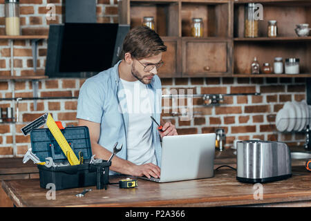 young man in eyeglasses using laptop while repairing toaster at home Stock Photo