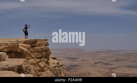 Young man standing on cliff edge and taking panoramic photo of the desert on his phone Stock Photo