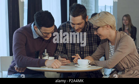 Group of young creative people wearing business casual clothes collaborating at meeting table and discussing work, using smartphone Stock Photo