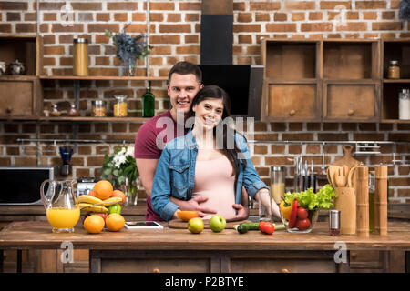 beautiful young pregnant couple hugging and smiling at camera while cooking together Stock Photo