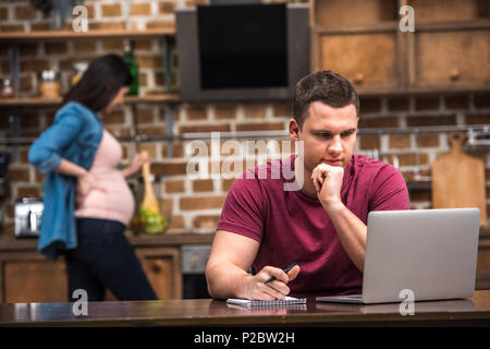 focused young man using laptop and taking notes while pregnant wife cooking behind Stock Photo