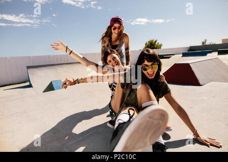 Three female friends playing with skateboard at the skate park. One woman pushing her friends from behind having fun and laughing. Stock Photo