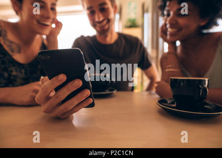 Three young people looking at mobile phone and smiling while sitting at restaurant table. Man showing something interesting to female friends during m Stock Photo
